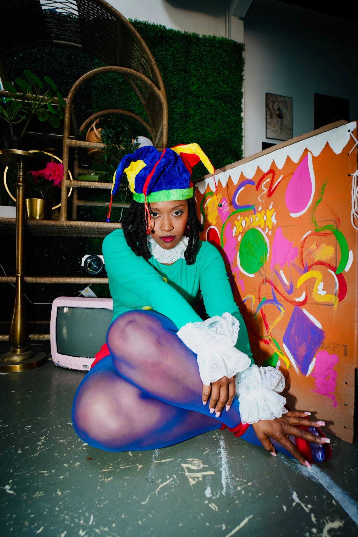 A woman in a colourful jester hat, turquoise top, white ruffled cuffs, and purple tights seated beside vivid artwork